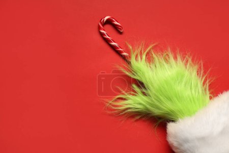 Téléchargez les photos : Main poilue verte de créature en costume de Père Noël avec canne à bonbons sur fond rouge - en image libre de droit