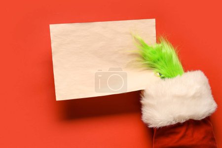 Photo for Green hairy hand of creature in Santa costume with blank paper sheet on red background - Royalty Free Image