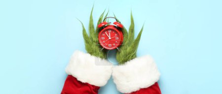Photo for Green hairy hands of creature in Santa costume holding alarm clock on light blue background, top view. Christmas countdown - Royalty Free Image