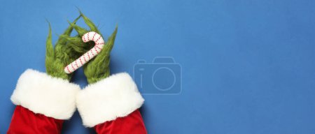 Téléchargez les photos : Mains poilues vertes de créature en costume de Père Noël tenant canne à bonbons de Noël sur fond bleu avec espace pour le texte - en image libre de droit