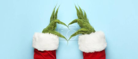 Green hairy hands of creature in Santa costume making heart on light blue background