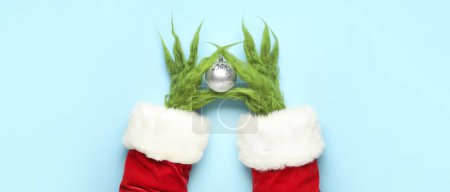 Téléchargez les photos : Mains poilues vertes de créature en costume de Père Noël tenant boule de Noël sur fond bleu clair - en image libre de droit