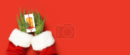 Téléchargez les photos : Mains poilues vertes de créature en costume de Père Noël tenant cadeau de Noël sur fond rouge avec espace pour le texte, vue du dessus - en image libre de droit