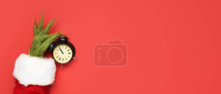 Photo for Green hairy hand of creature in Santa costume holding alarm clock on red background with space for text. Christmas countdown - Royalty Free Image