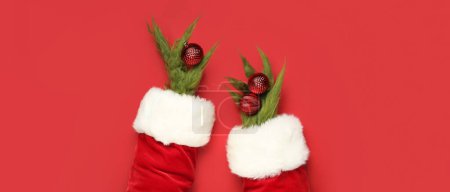 Téléchargez les photos : Mains poilues vertes de créature en costume de Père Noël tenant des boules de Noël sur fond rouge - en image libre de droit