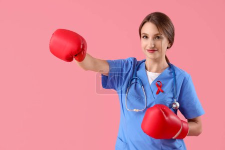 Foto de Doctora joven con cinta roja y guantes de boxeo sobre fondo rosa. Concepto del Día Mundial del SIDA - Imagen libre de derechos