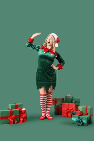 Foto de Mujer joven y feliz vestida de elfo con regalos de Navidad saludando mano sobre fondo verde - Imagen libre de derechos