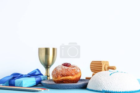 Photo for Gift box with dreidels, candles, cup and tasty donut on blue table near white wall. Hanukkah celebration - Royalty Free Image