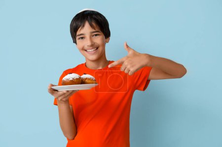 Photo for Little boy in kipa pointing at plate with tasty donuts on blue background. Hanukkah celebration - Royalty Free Image