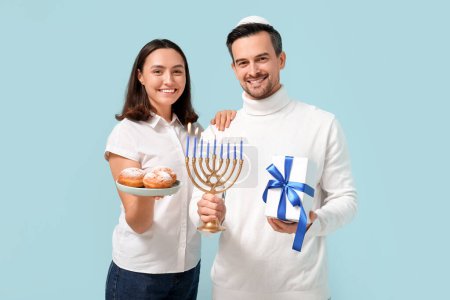 Téléchargez les photos : Joli couple avec menorah, sufganiyots et coffret cadeau sur fond bleu. Hanoukka célébration - en image libre de droit