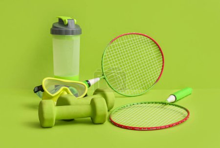 Photo for Dumbbells with sports bottle, swimming goggles and badminton rackets on green background - Royalty Free Image