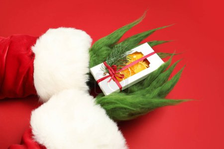Téléchargez les photos : Mains poilues vertes de créature en costume de Père Noël avec boîte d'oranges séchées sur fond rouge - en image libre de droit