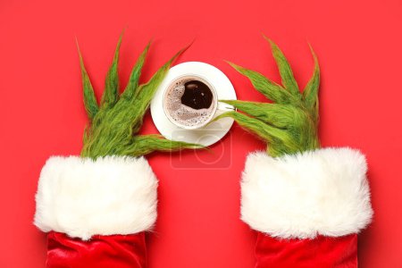 Verde manos peludas de criatura en traje de Santa con taza de café sobre fondo rojo