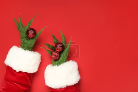 Téléchargez les photos : Mains poilues de créature verte en costume de Père Noël avec des boules de Noël sur fond rouge - en image libre de droit