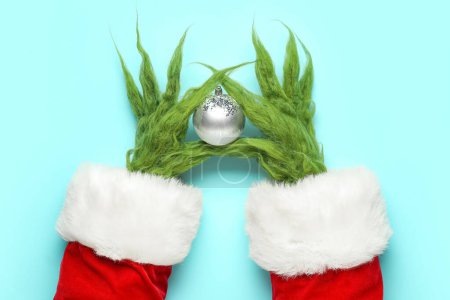 Photo for Green hairy hands of creature in Santa costume with Christmas ball on blue background - Royalty Free Image