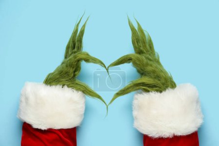 Green hairy hands of creature in Santa costume showing heart gesture on blue background