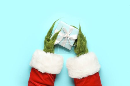 Photo for Green hairy hands of creature in Santa costume with gift box on blue background - Royalty Free Image