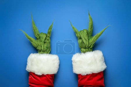 Téléchargez les photos : Mains poilues vertes de créature en costume de Père Noël montrant "cornes du diable" geste sur fond bleu - en image libre de droit