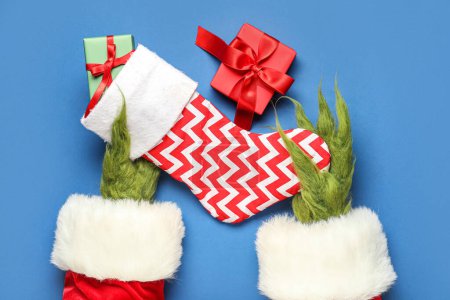 Photo for Green hairy hands of creature in Santa costume with Christmas sock and gift boxes on blue background - Royalty Free Image