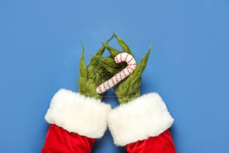 Photo for Green hairy hands of creature in Santa costume with decorative candy cane on blue background - Royalty Free Image