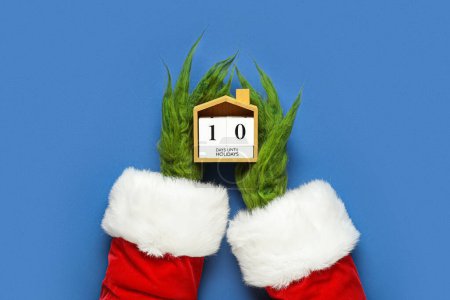 Photo for Green hairy hands of creature in Santa costume with countdown calendar on blue background - Royalty Free Image