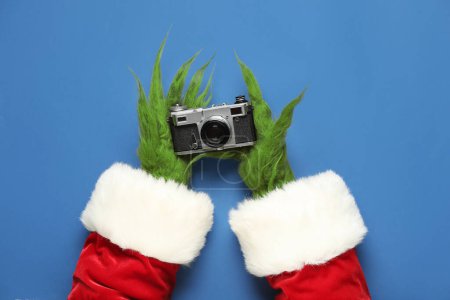 Téléchargez les photos : Mains poilues vertes de créature en costume de Père Noël avec caméra sur fond bleu - en image libre de droit
