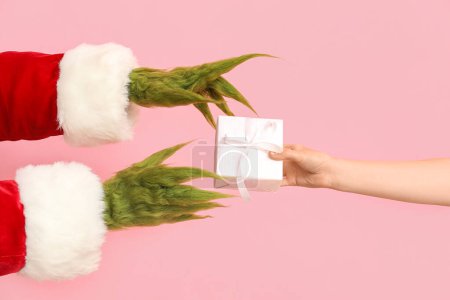 Photo for Green hairy creature in Santa costume and female hand with gift box on pink background - Royalty Free Image