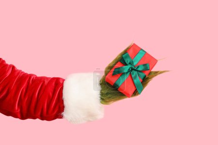Téléchargez les photos : Main poilue verte de créature en costume de Père Noël avec boîte cadeau sur fond rose - en image libre de droit