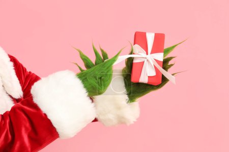 Green hairy hands of creature in Santa costume with gift boxes on pink background