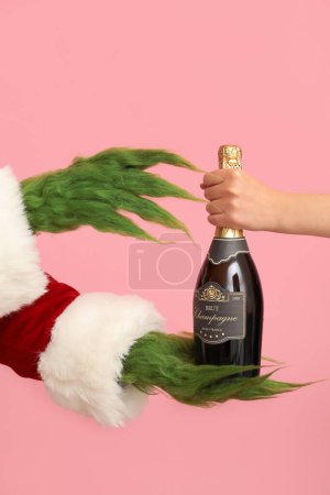 Téléchargez les photos : Créature poilue verte en costume de Père Noël et main féminine avec bouteille de champagne sur fond rose - en image libre de droit