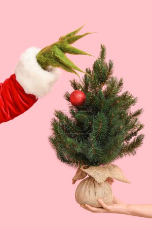 Green hairy creature in Santa costume and female hand with Christmas tree on pink background