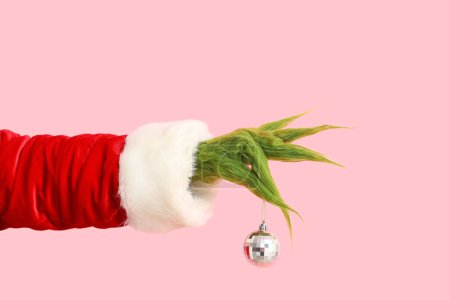 Green hairy hand of creature in Santa costume with Christmas ball on pink background