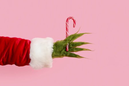 Téléchargez les photos : Main poilue verte de créature en costume de Père Noël avec canne à bonbons sur fond rose - en image libre de droit