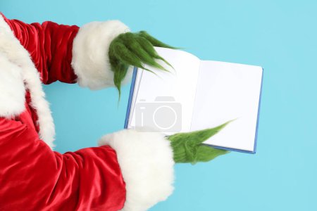 Téléchargez les photos : Mains poilues vertes de créature en costume de Père Noël avec carnet vierge sur fond bleu - en image libre de droit