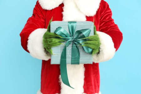 Foto de Criatura peluda verde en traje de Santa con caja de regalo sobre fondo azul, primer plano - Imagen libre de derechos