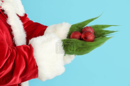 Photo for Green hairy hands of creature in Santa costume with Christmas balls on blue background - Royalty Free Image