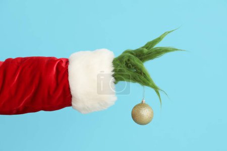 Téléchargez les photos : Main poilue verte de créature en costume de Père Noël avec boule de Noël sur fond bleu - en image libre de droit