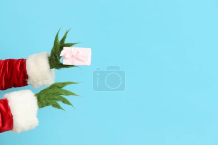 Téléchargez les photos : Mains poilues vertes de créature en costume de Père Noël avec boîte cadeau sur fond bleu - en image libre de droit