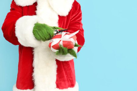 Photo for Green hairy creature in Santa costume with gift box and scissors on blue background, closeup - Royalty Free Image