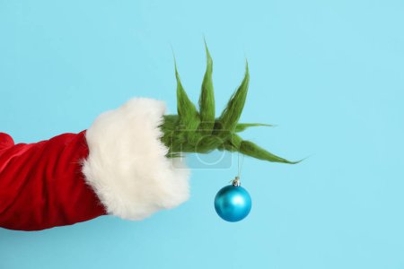Verde mano peluda de la criatura en traje de Santa con bola de Navidad sobre fondo azul