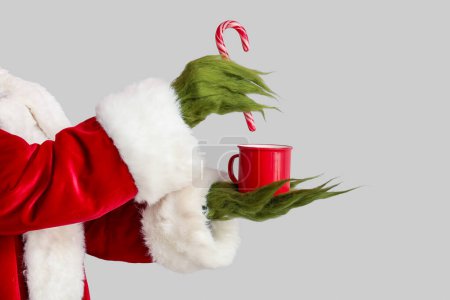 Criatura peluda verde en traje de Santa con taza de café y bastón de caramelo sobre fondo gris