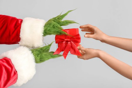 Photo for Green hairy creature in Santa costume and female hands with gift box on grey background - Royalty Free Image