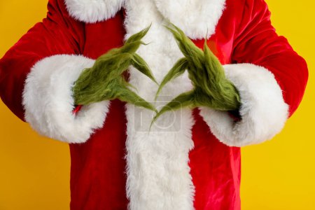 Téléchargez les photos : Créature poilue verte en costume de Père Noël montrant un geste de coeur sur fond jaune, gros plan - en image libre de droit