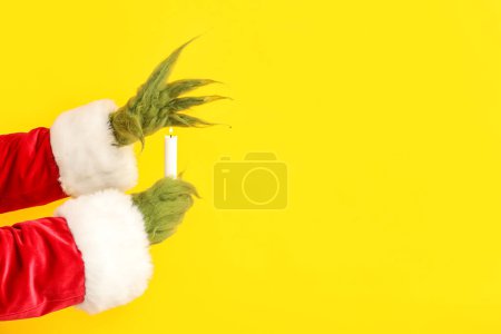 Téléchargez les photos : Mains poilues vertes de créature en costume de Père Noël avec bougie sur fond jaune - en image libre de droit