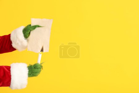 Photo for Green hairy hands of creature in Santa costume holding candle and burning paper sheet on yellow background - Royalty Free Image