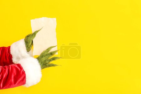 Photo for Green hairy hands of creature in Santa costume with blank paper sheet on yellow background - Royalty Free Image