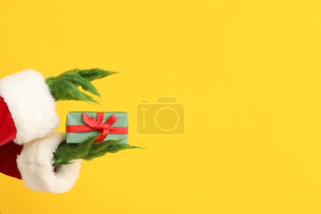 Téléchargez les photos : Mains poilues vertes de créature en costume de Père Noël avec boîte cadeau sur fond jaune - en image libre de droit