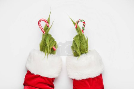 Téléchargez les photos : Mains poilues vertes de créature en costume de Père Noël avec des cannes à bonbons sur fond blanc - en image libre de droit