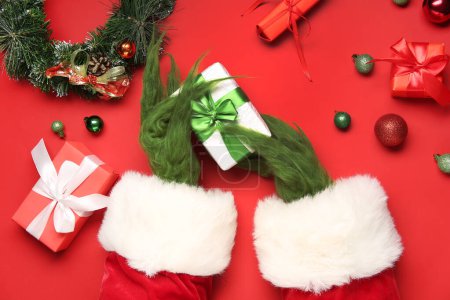 Téléchargez les photos : Mains poilues vertes de créature en costume de Père Noël avec des boîtes-cadeaux et des décorations de Noël sur fond rouge - en image libre de droit
