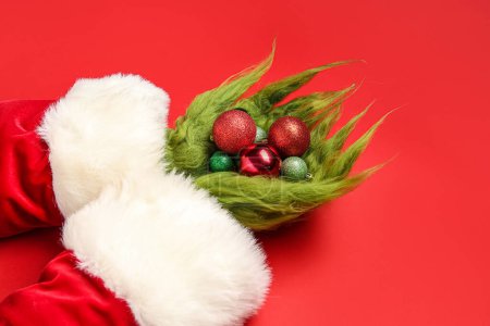 Téléchargez les photos : Mains poilues de créature verte en costume de Père Noël avec des boules de Noël sur fond rouge - en image libre de droit